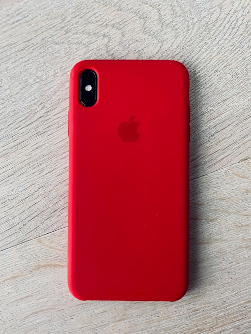 A smartphone with a red protective case resting on a light wooden surface. The case features a brand logo subtly embossed in the center. The camera lenses are visible on the top left corner of the case.