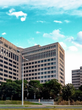 A large, modern office building with rectangular windows dominates the scene. The structure is surrounded by lush green trees and a neatly maintained lawn. A clear blue sky with scattered clouds serves as the backdrop.