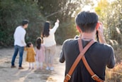 A person wearing a black shirt with a brown strap is taking a photo of a family in an outdoor setting. The family consists of two adults and two children, standing together on a dirt path with trees and greenery in the background. One of the adults is pointing towards something in the distance.