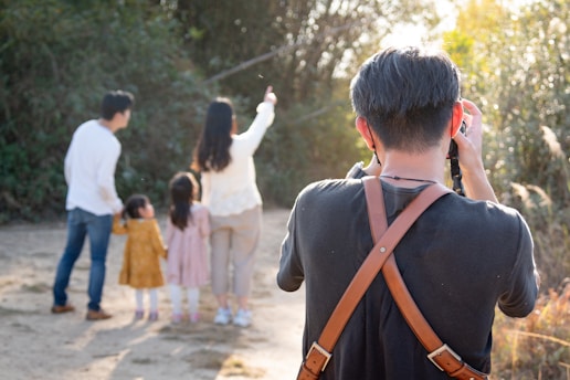A person wearing a black shirt with a brown strap is taking a photo of a family in an outdoor setting. The family consists of two adults and two children, standing together on a dirt path with trees and greenery in the background. One of the adults is pointing towards something in the distance.