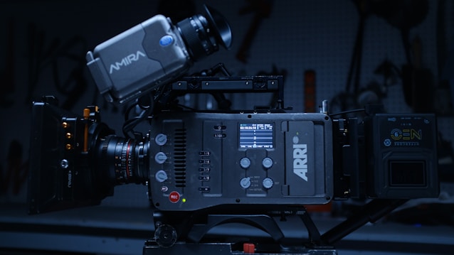 A professional-grade ARRI camera with an Amira viewfinder and various control buttons and dials is mounted on a tripod or support structure. The background is dimly lit, with indistinct shapes and tools visible, suggesting a workshop or studio environment.