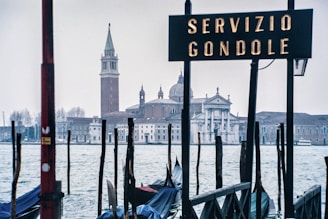 A scene depicting a gondola service area with a sign reading 'Servizio Gondole' in front of a historical building across the water. Numerous poles and boats are visible in the foreground with a church and bell tower in the background.