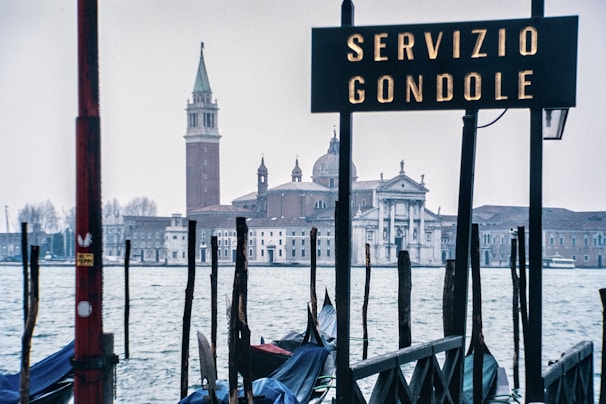 A scene depicting a gondola service area with a sign reading 'Servizio Gondole' in front of a historical building across the water. Numerous poles and boats are visible in the foreground with a church and bell tower in the background.