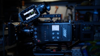 A professional digital film camera, specifically an ARRI model, with various controls and a display screen. It is set up on a tripod in a dimly lit environment with shelves and tools visible in the background.
