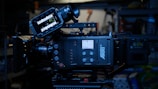 A professional digital film camera, specifically an ARRI model, with various controls and a display screen. It is set up on a tripod in a dimly lit environment with shelves and tools visible in the background.