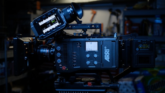 A professional digital film camera, specifically an ARRI model, with various controls and a display screen. It is set up on a tripod in a dimly lit environment with shelves and tools visible in the background.