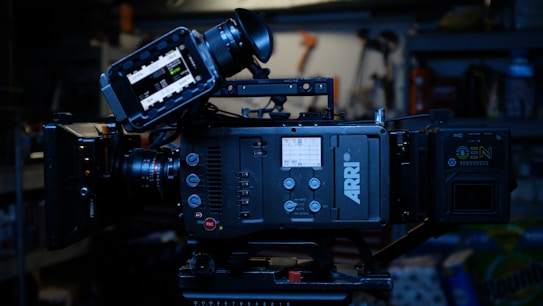 A professional digital film camera, specifically an ARRI model, with various controls and a display screen. It is set up on a tripod in a dimly lit environment with shelves and tools visible in the background.