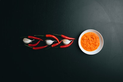 A black surface features an artistic arrangement of red chili peppers and garlic cloves on the left, leading to a white bowl filled with an orange, textured sauce on the right.