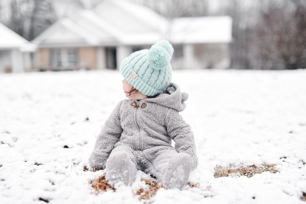 Kind in grauer Jacke und grüner Strickmütze sitzt tagsüber auf schneebedecktem Boden