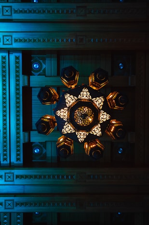 A geometric ceiling design with intricate wooden carvings and a central decorative chandelier featuring illuminated, ornate patterns.
