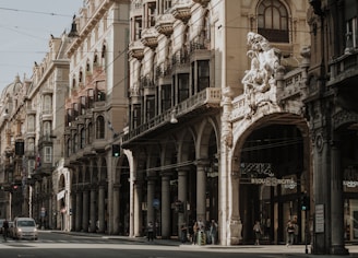 An ornate and historic street with intricate architecture featuring arched colonnades and elaborate sculptures. The buildings are multi-storied with decorative balconies and detailed facades. Pedestrians and cars are visible along the street, capturing the essence of a lively urban environment.