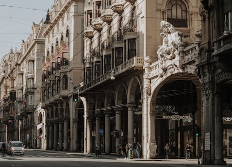 An ornate and historic street with intricate architecture featuring arched colonnades and elaborate sculptures. The buildings are multi-storied with decorative balconies and detailed facades. Pedestrians and cars are visible along the street, capturing the essence of a lively urban environment.