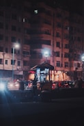 A nighttime street scene featuring a small food cart illuminated by bright lights. The vendor stands next to the cart, which is positioned on a sidewalk in front of a large residential building. Traffic is present in the background, with several cars visible under streetlights.