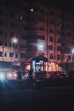A nighttime street scene featuring a small food cart illuminated by bright lights. The vendor stands next to the cart, which is positioned on a sidewalk in front of a large residential building. Traffic is present in the background, with several cars visible under streetlights.