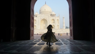 A silhouette of a person twirling inside an arched doorway frames the iconic Taj Mahal in the background. The architecture features intricate details with the white marble of the Taj Mahal glowing in the daylight. A few other people are visible in the distance, adding scale to the grand scene.