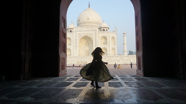 A silhouette of a person twirling inside an arched doorway frames the iconic Taj Mahal in the background. The architecture features intricate details with the white marble of the Taj Mahal glowing in the daylight. A few other people are visible in the distance, adding scale to the grand scene.