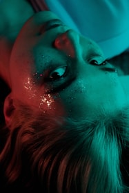 A person with glitter on their face lies on their back. The lighting casts a teal and reddish glow on their skin and hair, creating a surreal and dreamy atmosphere.