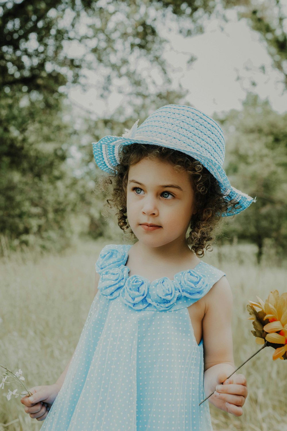 girl in blue dress and white hat holding yellow flower