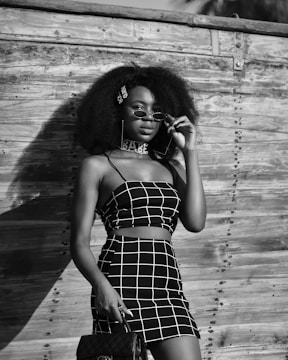 A person with a large, curly hairstyle wearing a black and white checkered outfit stands confidently against a rustic wooden background. They are holding sunglasses with one hand and a stylish handbag in the other.