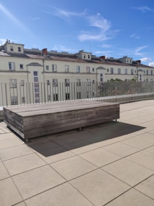 A rooftop terrace with light-colored tiles leads to a wooden rectangular bench. In the background, there is a series of apartment buildings with white walls and tiled roofs. The sky is bright and clear with scattered wispy clouds.