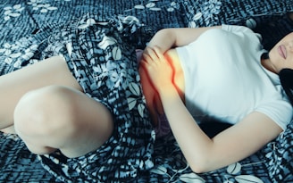 A person lies on a patterned bed, gently holding their abdomen. They are wearing a white top and the sheets have a blue and white floral design.