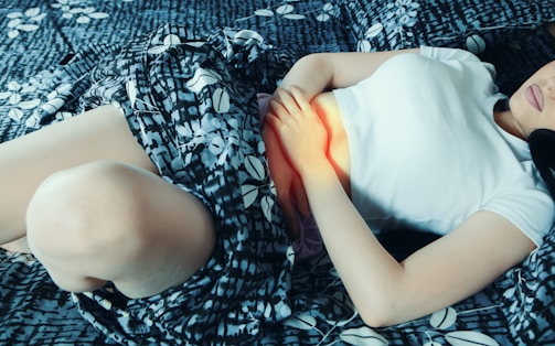 A person lies on a patterned bed, gently holding their abdomen. They are wearing a white top and the sheets have a blue and white floral design.