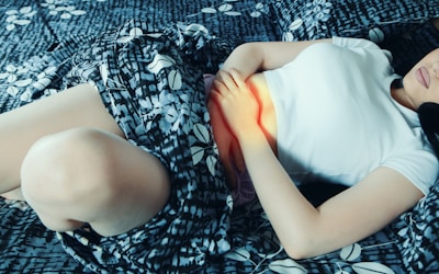 A person lies on a patterned bed, gently holding their abdomen. They are wearing a white top and the sheets have a blue and white floral design.