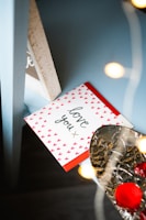A greeting card with 'love you' written on it is adorned with small red heart patterns. It rests on a blue surface beside a silver, textured object with red rose petals. Warm, soft lighting from out-of-focus lights creates a cozy ambiance.