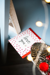 A greeting card with 'love you' written on it is adorned with small red heart patterns. It rests on a blue surface beside a silver, textured object with red rose petals. Warm, soft lighting from out-of-focus lights creates a cozy ambiance.