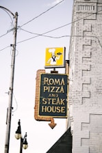 A vintage sign attached to a brick building advertises 'Roma Pizza and Steak House' with bold yellow lettering on a dark background. Above the sign, a depiction of a chef holding a pizza is displayed against a bright yellow background. Electrical wires and a streetlight are visible in the foreground, adding an urban, slightly nostalgic touch to the scene.