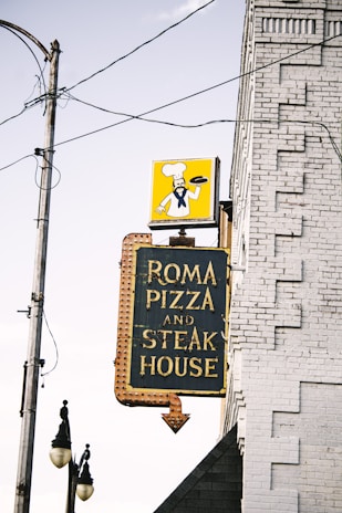A vintage sign attached to a brick building advertises 'Roma Pizza and Steak House' with bold yellow lettering on a dark background. Above the sign, a depiction of a chef holding a pizza is displayed against a bright yellow background. Electrical wires and a streetlight are visible in the foreground, adding an urban, slightly nostalgic touch to the scene.