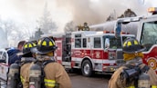 A group of firefighters in protective gear are managing a fire scene. They stand near a fire truck, which is actively spraying water. Smoke billows in the background, indicating a fire being tackled. The firefighters are wearing helmets and have equipment strapped to their backs.