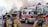 A group of firefighters in protective gear are managing a fire scene. They stand near a fire truck, which is actively spraying water. Smoke billows in the background, indicating a fire being tackled. The firefighters are wearing helmets and have equipment strapped to their backs.