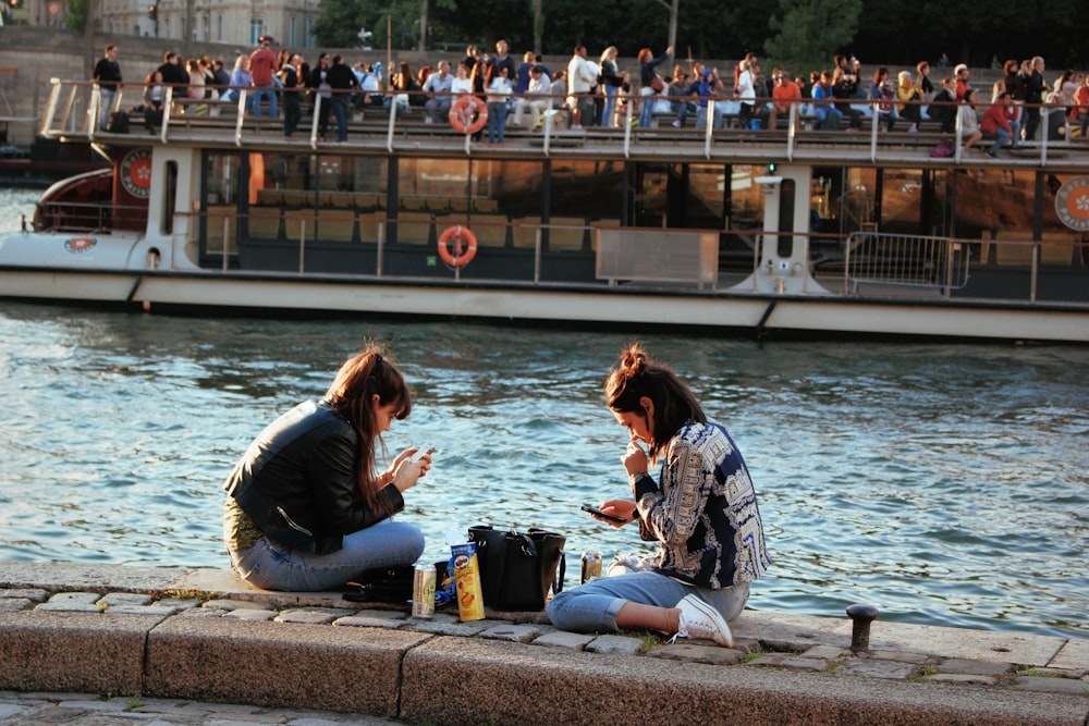 2 women sitting on concrete bench near body of water during daytime