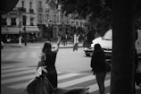 Two women stand at a street corner, one with her hand raised, possibly hailing a cab. A motorbike is parked nearby, and several buildings are visible across the street, along with a café. The scene depicts urban life in a city setting.