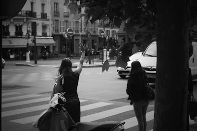 Two women stand at a street corner, one with her hand raised, possibly hailing a cab. A motorbike is parked nearby, and several buildings are visible across the street, along with a café. The scene depicts urban life in a city setting.