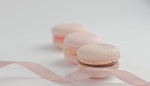 Three pastel-colored macarons are arranged in a soft-focus perspective on a light background. A pink ribbon is artfully draped in front of them, adding to the delicate and elegant presentation.