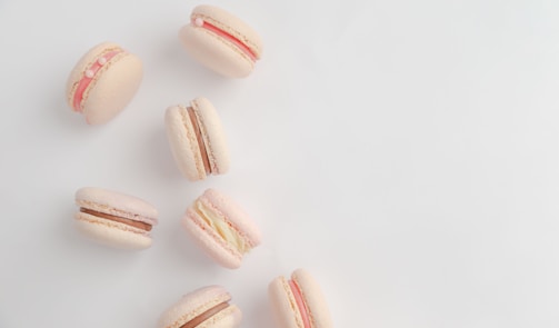 A group of pastel-colored macarons arranged on a light background. The macarons have various fillings, including pink, white, and chocolate. Some have decorative elements like small pearls.