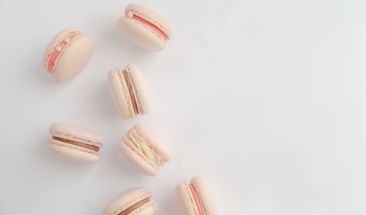 A group of pastel-colored macarons arranged on a light background. The macarons have various fillings, including pink, white, and chocolate. Some have decorative elements like small pearls.