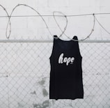 A black tank top with the word 'hope' printed on it is hanging on a chain link fence with barbed wire. The background is a white painted wall with some weathering visible.