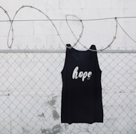 A black tank top with the word 'hope' printed on it is hanging on a chain link fence with barbed wire. The background is a white painted wall with some weathering visible.