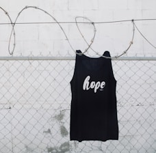 A black tank top with the word 'hope' printed on it is hanging on a chain link fence with barbed wire. The background is a white painted wall with some weathering visible.
