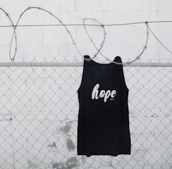 A black tank top with the word 'hope' printed on it is hanging on a chain link fence with barbed wire. The background is a white painted wall with some weathering visible.