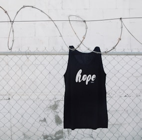 A black tank top with the word 'hope' printed on it is hanging on a chain link fence with barbed wire. The background is a white painted wall with some weathering visible.