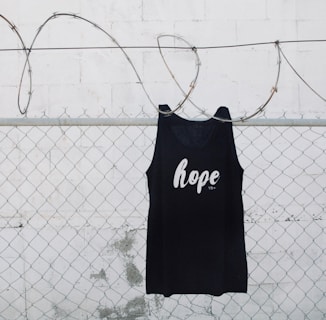 A black tank top with the word 'hope' printed on it is hanging on a chain link fence with barbed wire. The background is a white painted wall with some weathering visible.