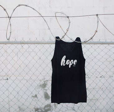 A black tank top with the word 'hope' printed on it is hanging on a chain link fence with barbed wire. The background is a white painted wall with some weathering visible.