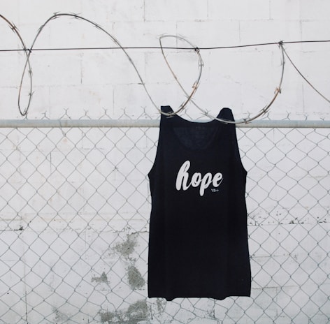 A black tank top with the word 'hope' printed on it is hanging on a chain link fence with barbed wire. The background is a white painted wall with some weathering visible.
