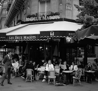 A bustling Parisian café scene with multiple people seated at outdoor tables, engaged in conversation and dining. The café, Les Deux Magots, features classic bistro chairs and tables. The atmosphere is lively and social, with a man in a suit walking by in the foreground.