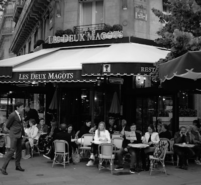 A bustling Parisian café scene with multiple people seated at outdoor tables, engaged in conversation and dining. The café, Les Deux Magots, features classic bistro chairs and tables. The atmosphere is lively and social, with a man in a suit walking by in the foreground.
