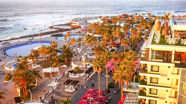 A tropical coastal resort with numerous palm trees, multiple swimming pools, and people walking along pathways. The image includes a large building with balconies and outdoor seating areas on the right. The ocean with waves crashing against rocks is visible in the background.